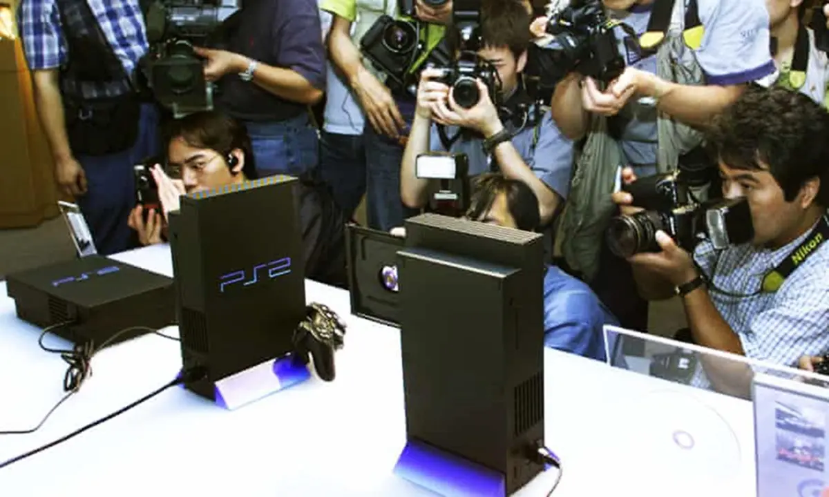 Evento de anuncio do PlayStation 2, com consoles em uma bancada branca e vários fotógrafos tirando fotos.