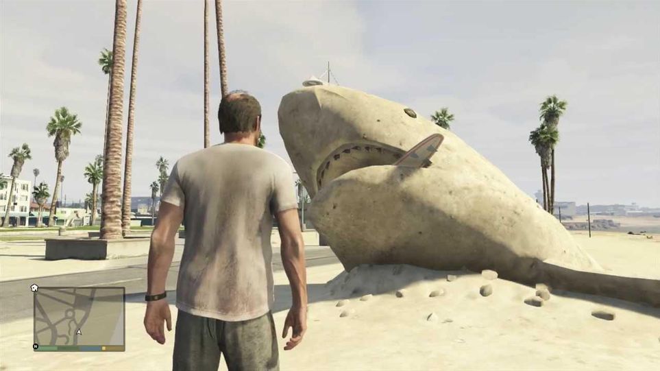 Trevor, personagem do jogo GTA V, encarando um tubarão feito de areia da praia.