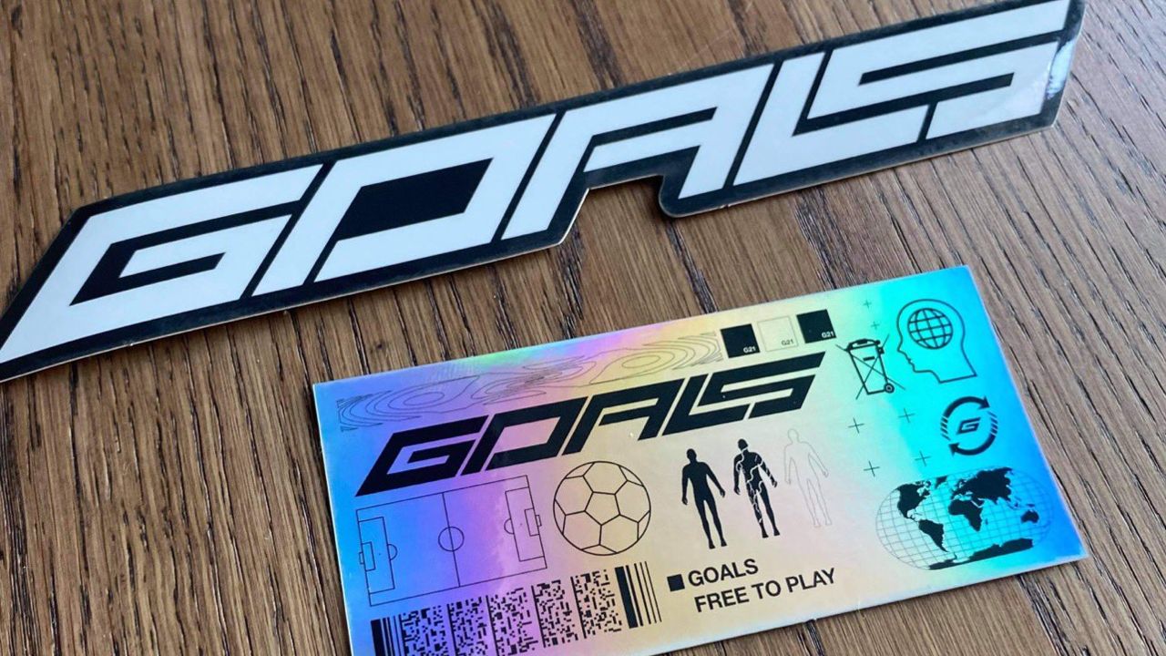 Imagem de capa da logo do jogo GOALS em cima de uma madeira e um cartão brilhante embaixo com referências ao futebol