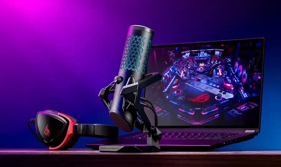 Foto do microfone preto em uma mesa, ao lado de um notebook e headset da Asus.